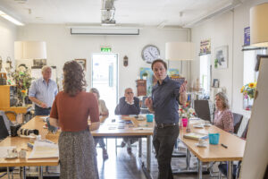 een groep mensen zit om tafel, en een man en vrouw presenteren iets op een flipover