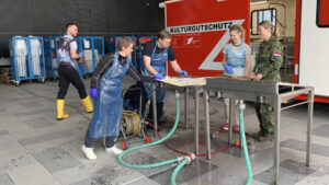 3 personen zijn bezig met een object op een werkbank onder leiding van iemand van defensie.