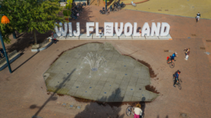 Luchtfoto van ''WIJ FLEVOLAND'' letters en betegeling in de vorm van de noordoostpolder waaruit een fontein sproeit.